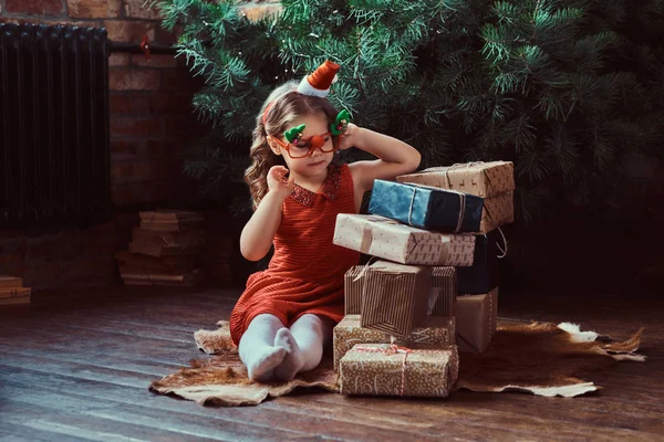 Portrait of a cute girl with blonde curly hair wearing a red dress and little Santas hat sitting on a floor surrounded by gifts — Stock Photo, Image