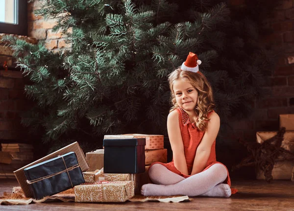 Retrato de uma menina bonita com cabelo encaracolado loiro vestindo um vestido vermelho e chapéu pequeno Santas sentado em um chão cercado por presentes — Fotografia de Stock