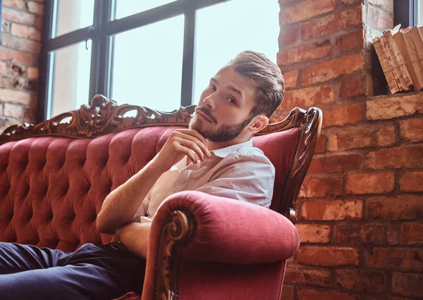 Retrato de un joven guapo con una elegante barba y cabello elegantemente vestido sentado en un sofá rojo vintage —  Fotos de Stock