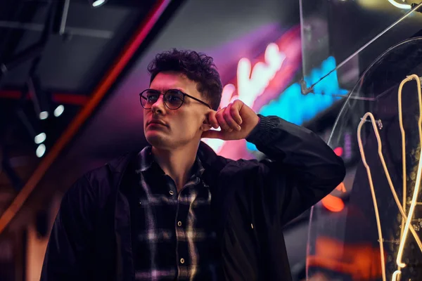 A pensive young man leaning on an illuminated signboard in the street at night. — Stock Photo, Image