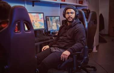 Afro-Amerikan sakallı adam hoodie ve oyun bir sandalyede oturan ve bir kamera bir oyun kulüp veya internet kafede bakarak kap giymiş.