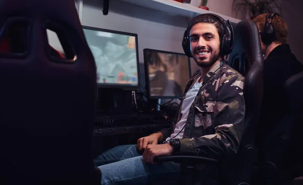 Um jovem indiano alegre vestindo uma camisa militar sentado em uma cadeira de jogador e olhando para uma câmera em um clube de jogos ou café na internet . — Fotografia de Stock