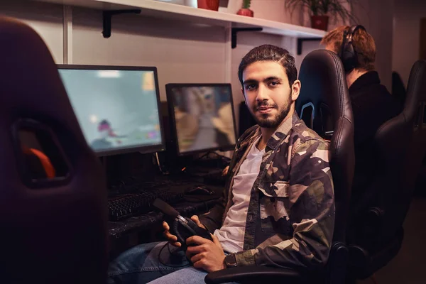 Um jovem indiano vestindo uma camisa militar sentado em uma cadeira de jogador e olhando para uma câmera em um clube de jogos ou internet café . — Fotografia de Stock