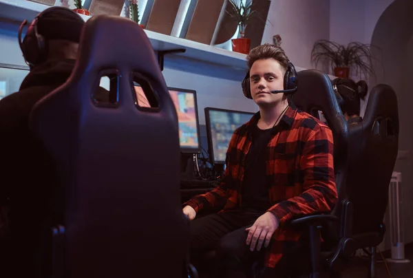 A young stylish guy wearing a checkered shirt and headset sitting on a gamer chair and looking at a camera in a gaming club or internet cafe. — Stock Photo, Image