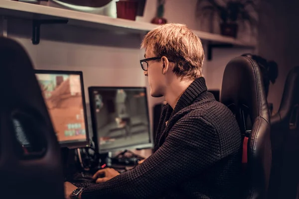 Un joven vistiendo un suéter y gafas sentado en una silla de jugador y jugar videojuegos en un club de juegos o cibercafé . —  Fotos de Stock