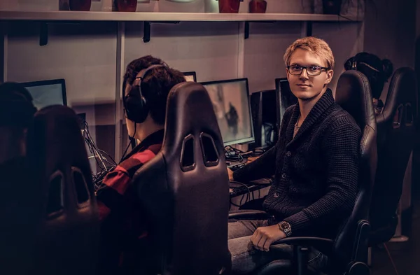 Um jovem jogador inteligente usando uma camisola e óculos sentados em uma cadeira de jogador e olhando para uma câmera em um clube de jogos ou café na internet . — Fotografia de Stock