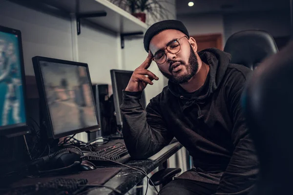 Cansado jogador barbudo afro-americano vestindo capuz e boné adormeceu sentado na mesa de computador em um clube de jogos ou internet café . — Fotografia de Stock