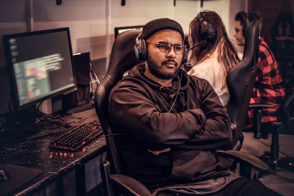 African-American bearded guy wearing hoodie and cap with her arms crossed sitting on a gamer chair in a gaming club or internet cafe. — Stock Photo, Image