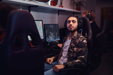 Genç bir Hintli adam oyun bir sandalyede oturan ve bir kamera bir oyun kulüp veya internet kafede bakarak askeri bir gömlek giymiş.