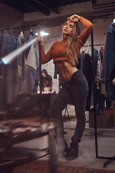 Уставшая девушка с голым животом и расстегнутыми штанами, опирающаяся на стойку с одеждой в примерочной магазина одежды . — стоковое фото