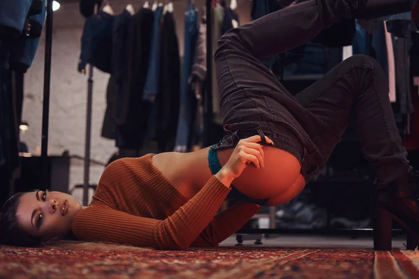 Ein schönes Mädchen, das verführerisch die Hosen hochzieht, während es in der Umkleidekabine eines Bekleidungsgeschäfts auf dem Boden liegt. — Stockfoto