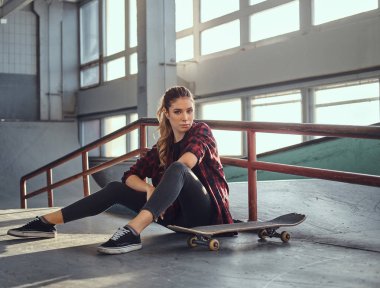 Skatepark kapalı bir eziyet demiryolu yanında otururken bir kaykay tutan bir kareli gömlek giymiş güzel bir genç kız.