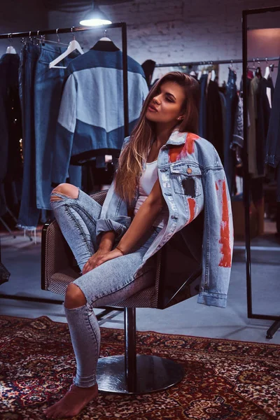 Una chica cansada vistiendo pantalones vaqueros y chaqueta de mezclilla en una silla en el probador de una tienda de ropa . — Foto de Stock