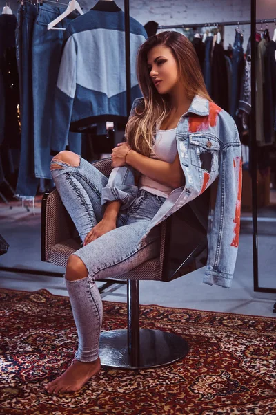 Una chica elegante con pantalones vaqueros y abrigo afligidos cubriendo su hombro sentado en una silla en el probador de una tienda de ropa . — Foto de Stock