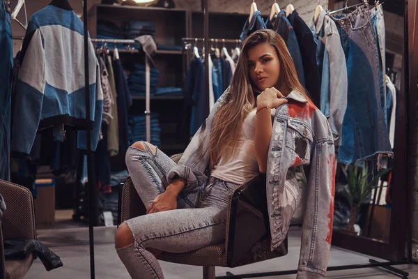 Elegantní dívka, která nosí zoufalý džíny a srst pokrývající rameno sedí na židli v zkušebna obchod s oblečením. — Stock fotografie