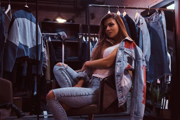 Uma menina elegante vestindo jeans angustiados e casaco cobrindo seu ombro sentado em uma cadeira no vestiário de uma loja de roupas . — Fotografia de Stock