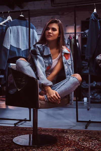 Стильная девушка в джинсах и джинсовой куртке, сидящая на стуле в примерочной в магазине одежды . — стоковое фото