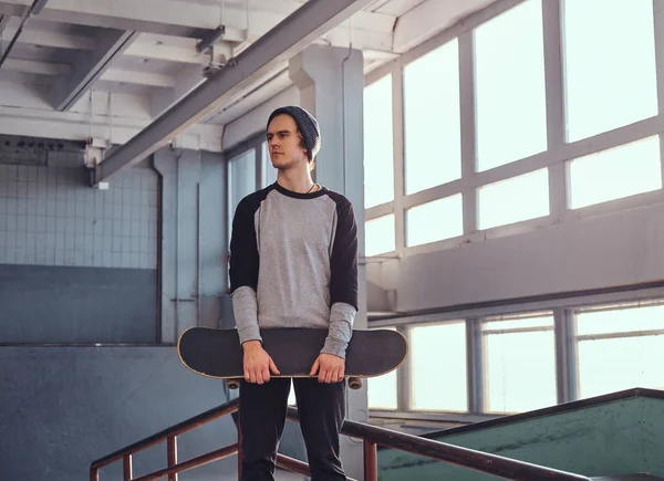 Jonge skateboarder permanent naast een spoor van de sleur in skatepark binnenshuis, zijn bestuur te houden en weg op zoek. — Stockfoto