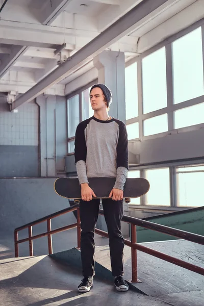 Молодой скейтбордист стоит рядом с молотым рельсом в скейтпарке в помещении, держа доску и отворачиваясь . — стоковое фото