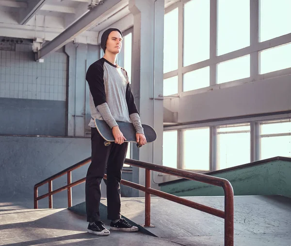 Молодой скейтбордист стоит рядом с молотым рельсом в скейтпарке в помещении, держа доску и отворачиваясь . — стоковое фото
