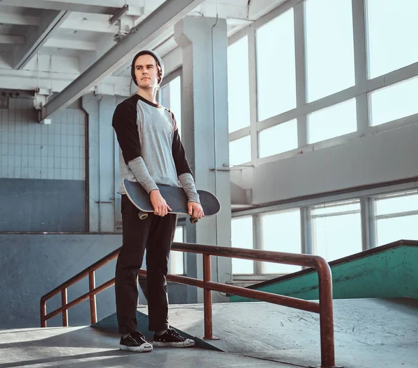 Молодий скейтбордист стоїть поруч з м'якою рейкою в скейтпарку в приміщенні, тримаючи свою дошку і дивлячись геть . — стокове фото