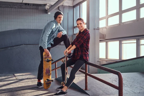 Alegre casal de jovens com skates vestindo roupas casuais posando ao lado de um trilho moagem no parque de skate dentro de casa . — Fotografia de Stock