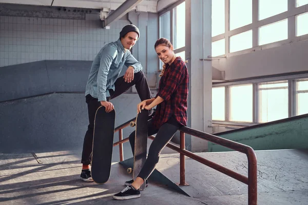 Alegre casal de jovens com skates vestindo roupas casuais posando ao lado de um trilho moagem no parque de skate dentro de casa . — Fotografia de Stock