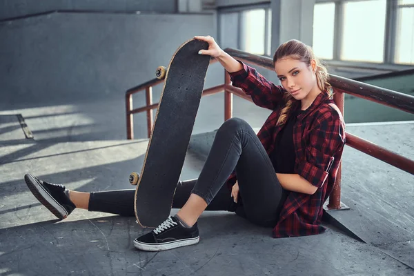 Ein schönes junges Mädchen im karierten Hemd hält ein Skateboard in der Hand, während sie im Skatepark neben einer Schleifschiene sitzt. — Stockfoto