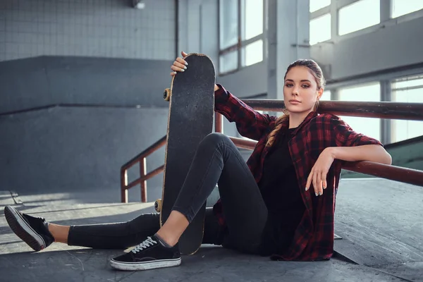 Una hermosa joven con una camisa a cuadros sosteniendo un monopatín mientras está sentada junto a un carril de molienda en el parque de skate en interiores . — Foto de Stock