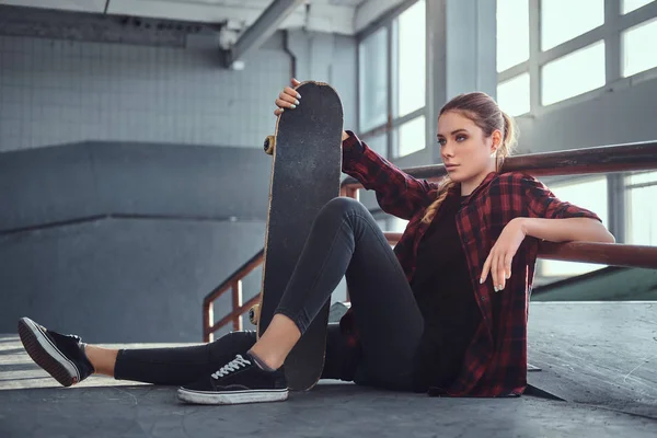 Красивая молодая девушка в клетчатой рубашке, держащая скейтборд, сидя рядом с молотым рельсом в скейтпарке в помещении . — стоковое фото