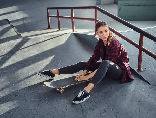 Een mooi jong meisje dragen een geruite shirt houden een skateboard zittend naast een spoor van de sleur in skatepark binnenshuis. — Stockfoto