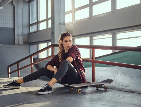 En vacker ung flicka som bär en rutig skjorta hålla en skateboard samtidigt sitter bredvid en grind järnväg i skatepark inomhus. — Stockfoto