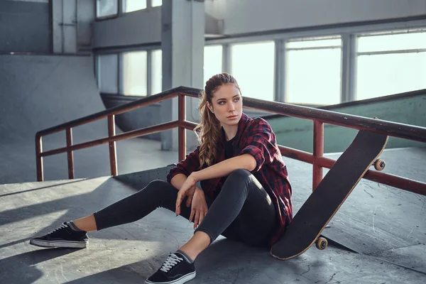 Una hermosa joven con una camisa a cuadros sosteniendo un monopatín mientras está sentada junto a un carril de molienda en el parque de skate en interiores . — Foto de Stock