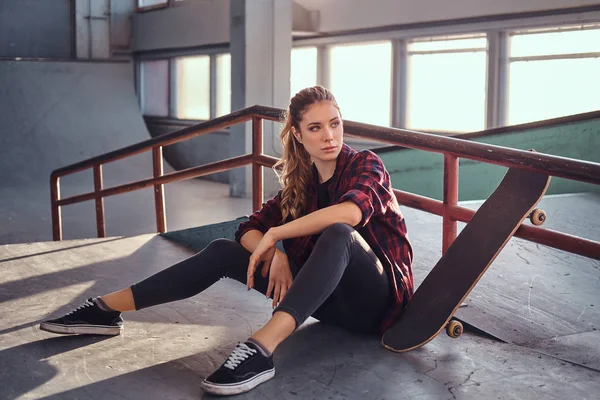 A beautiful young girl wearing a checkered shirt holding a skateboard while sitting next to a grind rail in skatepark indoors. — Stock Photo, Image