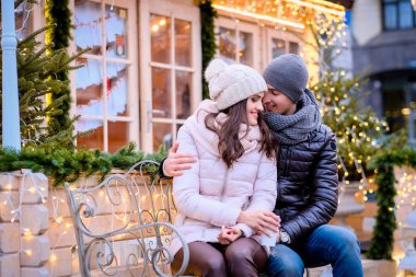 Sıcak giysiler giyiyor, bir tarihte birlikte vakit Noel zamanda dekore edilmiş bir sokakta bir bankta otururken sarılma zevk mutlu romantik çift