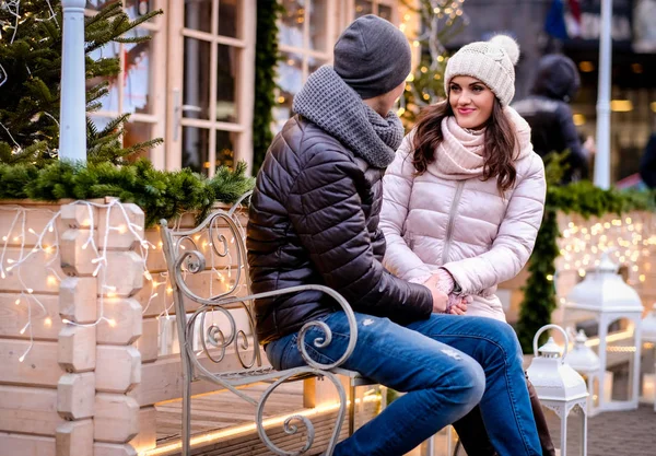 Casal romântico vestindo roupas quentes sentado em um banco na rua à noite decorado com belas luzes, falando e aquecendo com café na época do Natal ao ar livre — Fotografia de Stock