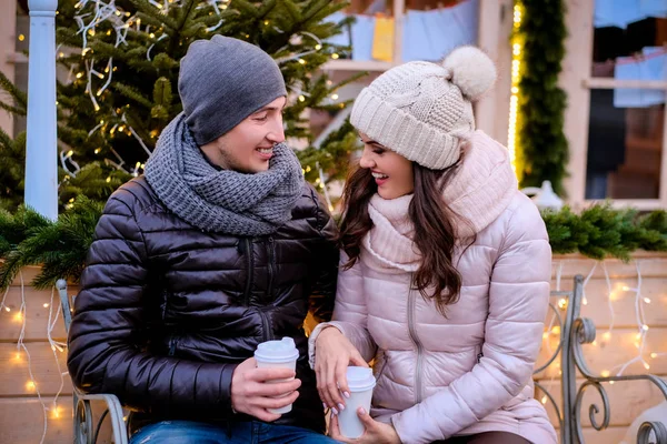 Casal romântico vestindo roupas quentes sentado em um banco na rua à noite decorado com belas luzes, falando e aquecendo com café na época do Natal ao ar livre — Fotografia de Stock