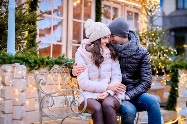 Casal romântico feliz vestindo roupas quentes, gostando de passar o tempo juntos em um encontro, abraçando enquanto sentado em um banco em uma rua decorada no tempo de Natal — Fotografia de Stock