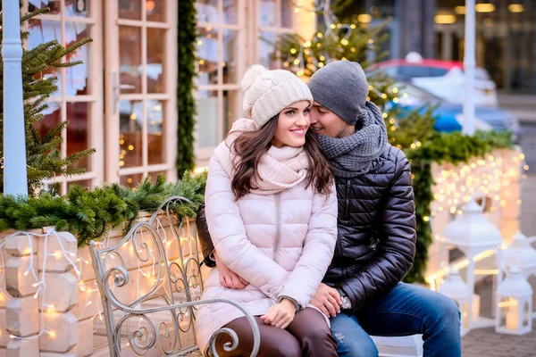 Happy romantic couple wearing warm clothes, enjoying spending time together on a date, hugging while sitting on a bench on a decorated street at Christmas time