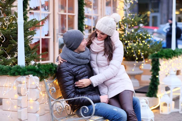 Casal romântico feliz vestindo roupas quentes, gostando de passar o tempo juntos em um encontro, abraçando enquanto sentado em um banco em uma rua decorada no tempo de Natal — Fotografia de Stock