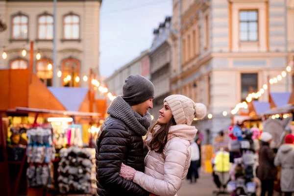 Una joven pareja romántica con ropa de invierno abrazándose mientras está de pie en la calle de la noche con la feria de Navidad — Foto de Stock