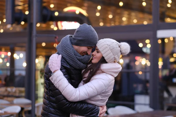 Una joven pareja romántica con ropa abrigada abrazándose juntos en la calle de la noche cerca de un café afuera en Navidad — Foto de Stock