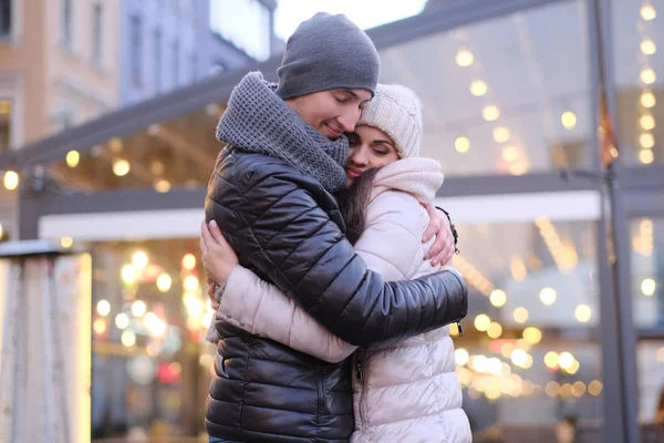 Um jovem casal romântico vestindo roupas quentes abraçando juntos na rua à noite perto de um café fora na época do Natal — Fotografia de Stock