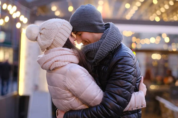Una joven pareja romántica con ropa abrigada abrazándose juntos en la calle de la noche cerca de un café afuera en Navidad —  Fotos de Stock