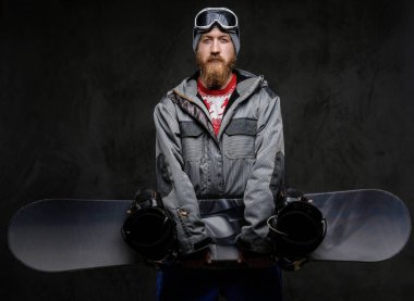 Tam bir ekipman snowboard tutarak ve karanlık dokulu bir arka plan üzerinde izole bir kamera bakarak giyen bir kırmızı sakallı yakışıklı adam.