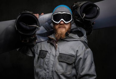 Acımasız adam karanlık dokulu bir arka plan üzerinde izole onun omzuna bir snowboard tutan bir tam ekipmanlı giyen kırmızı sakallı.
