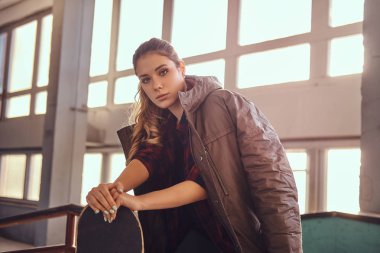 Bir kaykay üzerinde yaslanmış ve kameraya skatepark kapalı bir eziyet demiryolu üzerinde otururken görünümlü omuzlarını kapsayan bir kat ile güzel bir genç kız portresi.