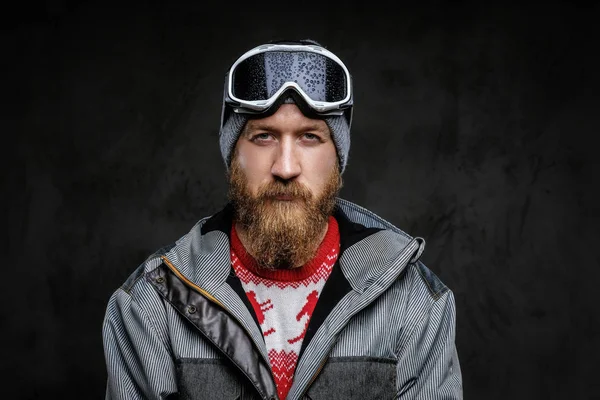 Confident man with a red beard wearing a full equipment for extreme snowboarding, looking at a camera with a serious look, isolated on a dark textured background. — Stock Photo, Image