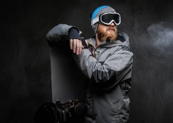 Redhead vousatý muž v kompletním vybavením pro extrémní snowboarding, opíraje se o snowboardu a koukal, izolované na tmavý podklad s texturou. — Stock fotografie