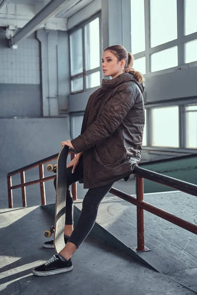 A beautiful young girl wearing a checkered shirt and coat with her skateboard sitting on a grind rail in skatepark indoors. — Stock Photo, Image